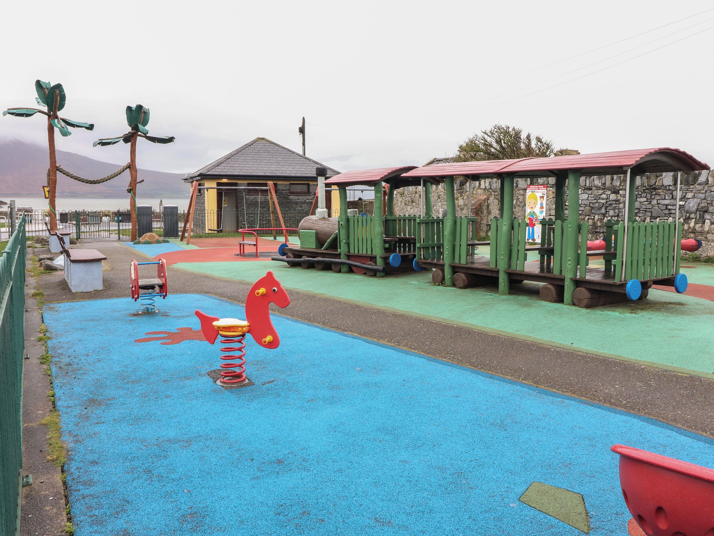 Une aire de jeux pour enfants avec une surface en caoutchouc bleue, des balançoires, une balançoire en forme de cheval rouge et un petit bâtiment avec un toit rouge.