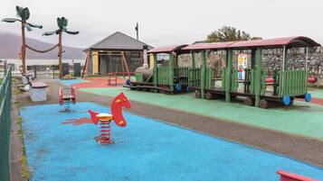 Une aire de jeux pour enfants avec une surface en caoutchouc bleue, des balançoires, une balançoire en forme de cheval rouge et un petit bâtiment avec un toit rouge.