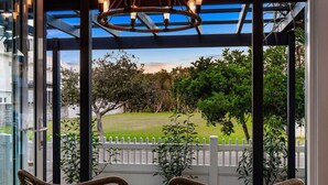 A covered outdoor seating area with wicker chairs, a white picket fence, and a view of a grassy park.