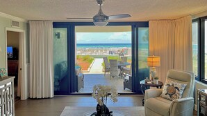 Una habitación con ventilador de techo, vista a la playa, una silla con almohada de tortuga y una mesa con lámpara y florero con orquídeas.