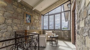 Una habitación con paredes de piedra, un ventanal grande, una silla blanca, una mesita pequeña y una escalera con barandilla de hierro forjado.