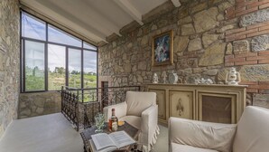 Una habitación acogedora con pared de piedra, un ventanal grande y vistas al campo. Hay una mesa con un libro, una botella y vasos.