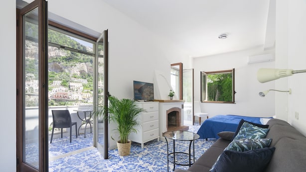 Une pièce avec un sol à motifs bleus, un canapé gris avec des coussins, une petite table ronde, une commode, un miroir et une vue sur un balcon agrémenté d’une plante.