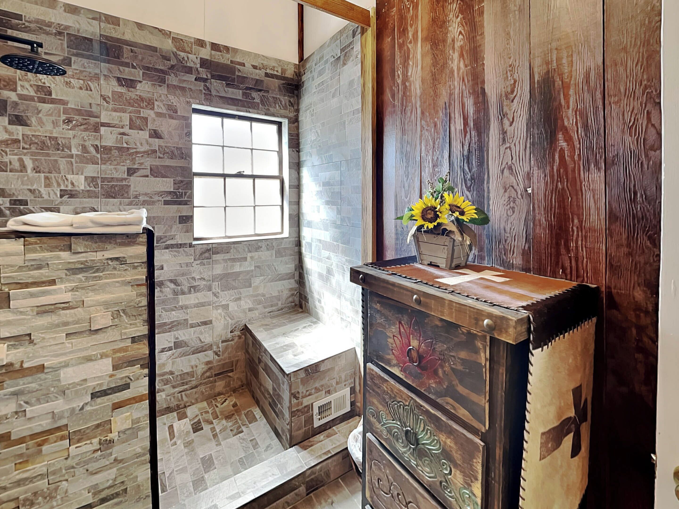 A bathroom with a stone wall, a window, a shower, and a wooden dresser with a vase of sunflowers.