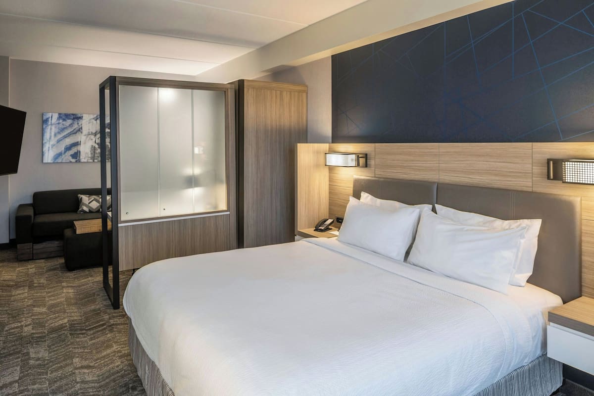 A modern hotel room with a large bed, a desk with a lamp, a wardrobe, and a wall-mounted TV.