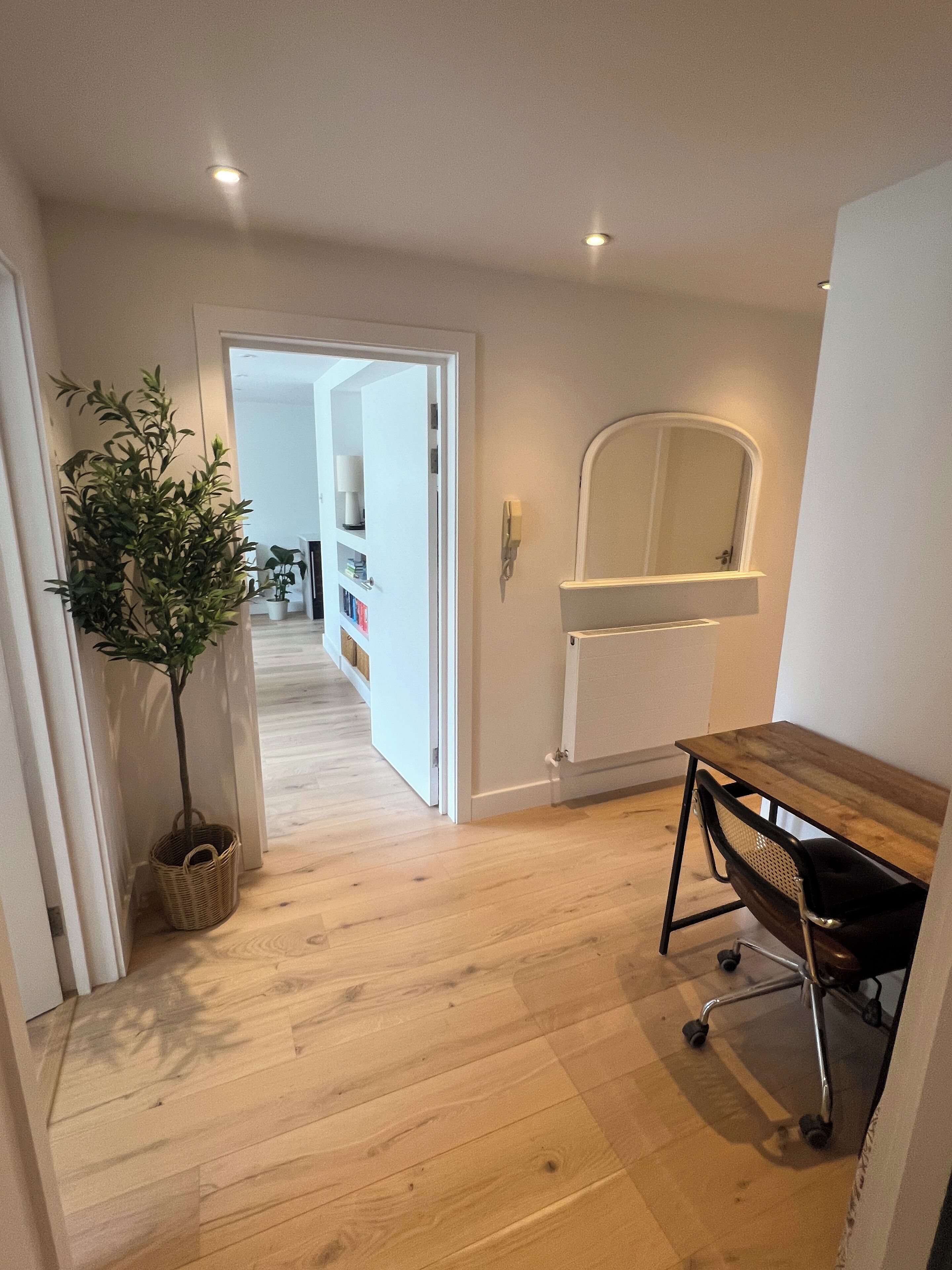 A hallway with wooden flooring, a potted plant, a desk with a chair, and a mirror on the wall.