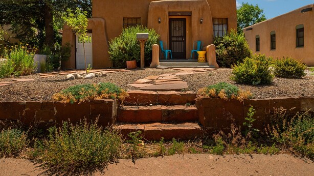 Un bâtiment traditionnel en adobe, doté d’une entrée principale, de marches menant à cette entrée et d’un petit jardin avec des plantes et un sentier.