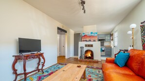 Un salon avec un téléviseur, une table basse en bois, un canapé orange, un tapis coloré, une cheminée et une cuisine en arrière-plan.