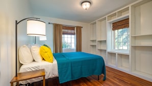 Une chambre à coucher comprenant un lit, une table de chevet, une lampe, une fenêtre avec des rideaux et des étagères intégrées.