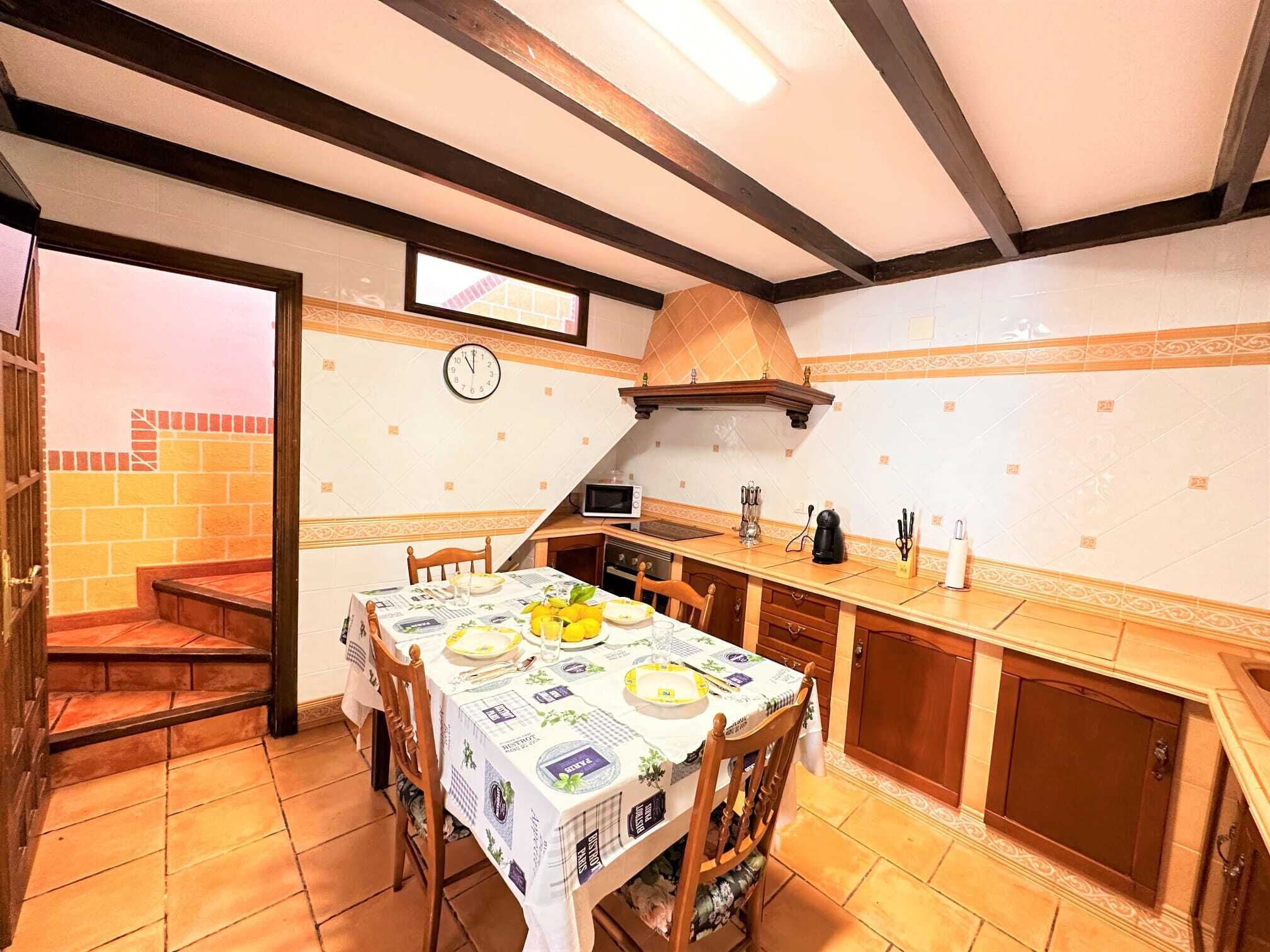 Une cuisine avec une table à manger pour quatre personnes, une horloge au mur et un escalier en bois menant à l’étage supérieur.