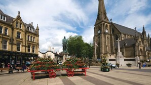 Una plaza histórica con una imponente torre de iglesia, bancos y arreglos florales.