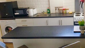 Cocina compacta con encimera negra, armarios blancos, microondas, cafetera, lavabo y una pequeña planta en maceta.