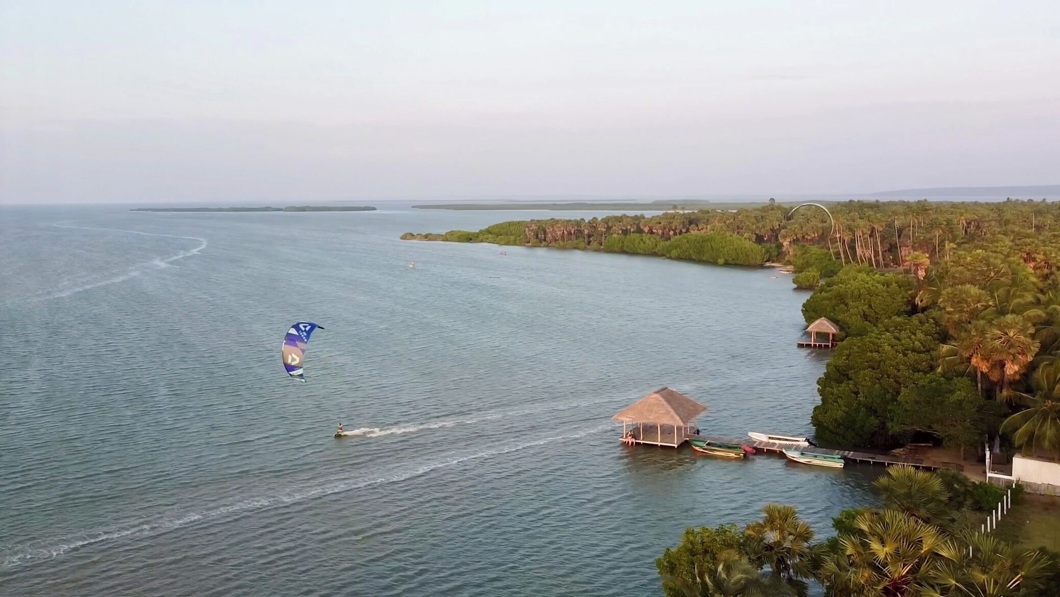 Een kustgebied met een parasail in het water, een klein steiger met een hutje, en weelderige begroeiing langs de oever.