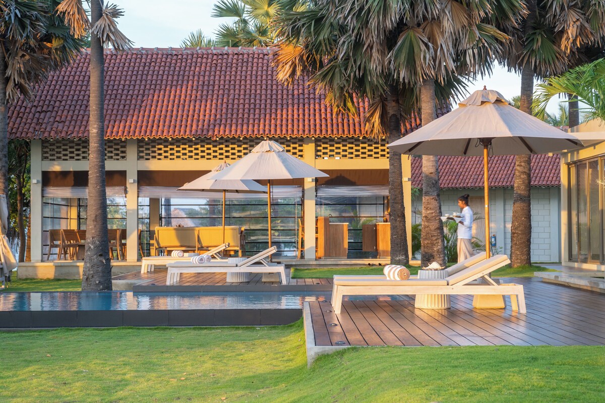 A modern outdoor pool area with lounge chairs and umbrellas, surrounded by palm trees and a building with a tiled roof.