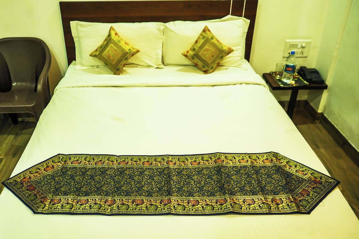 Habitación de hotel con cama, dos almohadas, una silla, mesita de noche con teléfono y botella de agua, y alfombra decorativa.