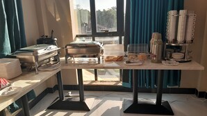 A buffet setup with food containers, plates, and utensils on a table near a window with curtains.