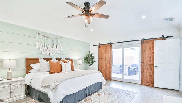 Een slaapkamer met een bed, een plafondventilator, een nachtkastje, een dressoir en een groot raam met uitzicht op de omgeving.