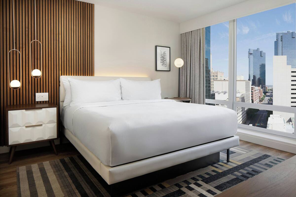 Een moderne hotelkamer met een groot bed, een wand van hout als accent, uitzicht over de stad, en een nachtkastje met een lamp.