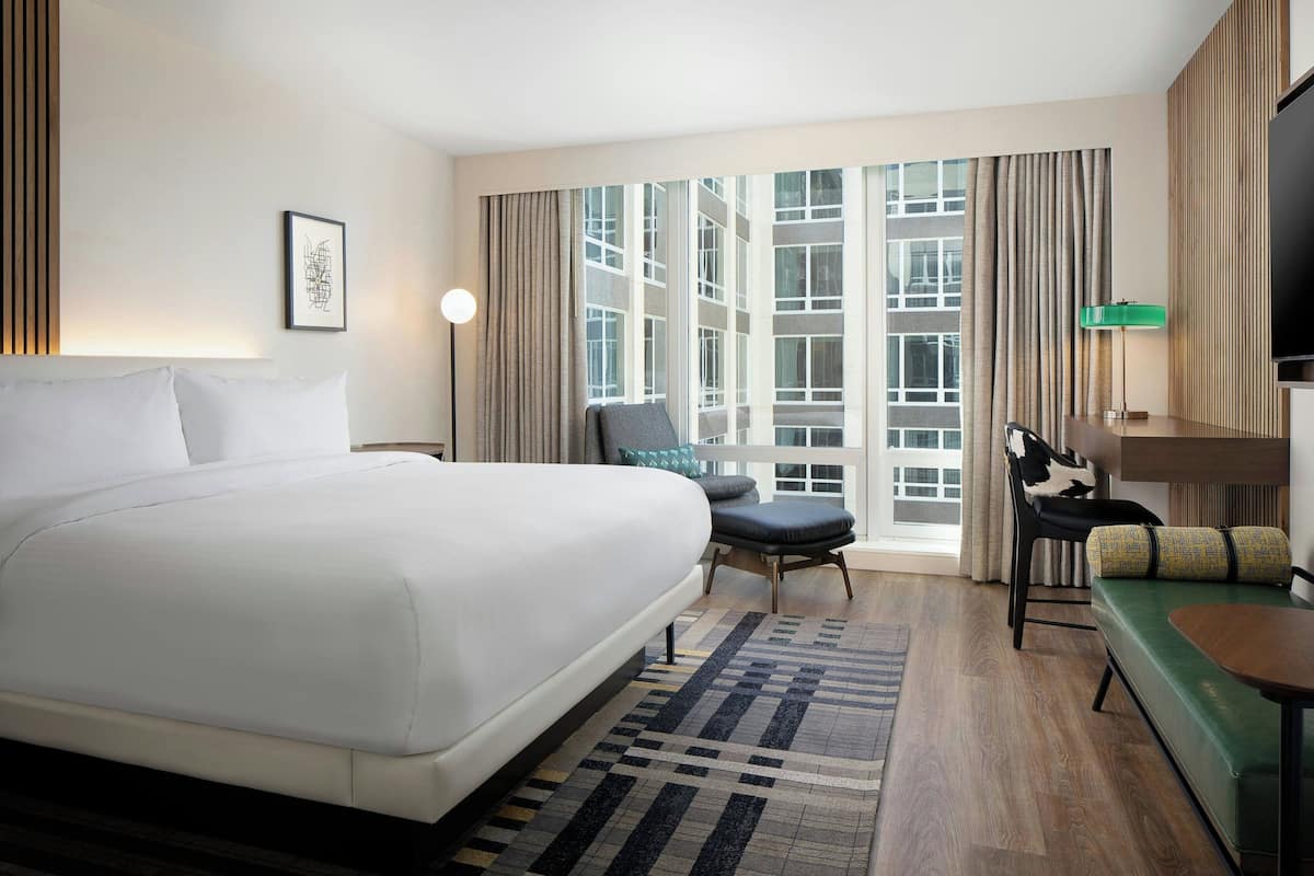 Hotelkamer met een groot bed, een bureau, een stoel en een bank. Er is een raam met gordijnen en uitzicht op gebouwen.