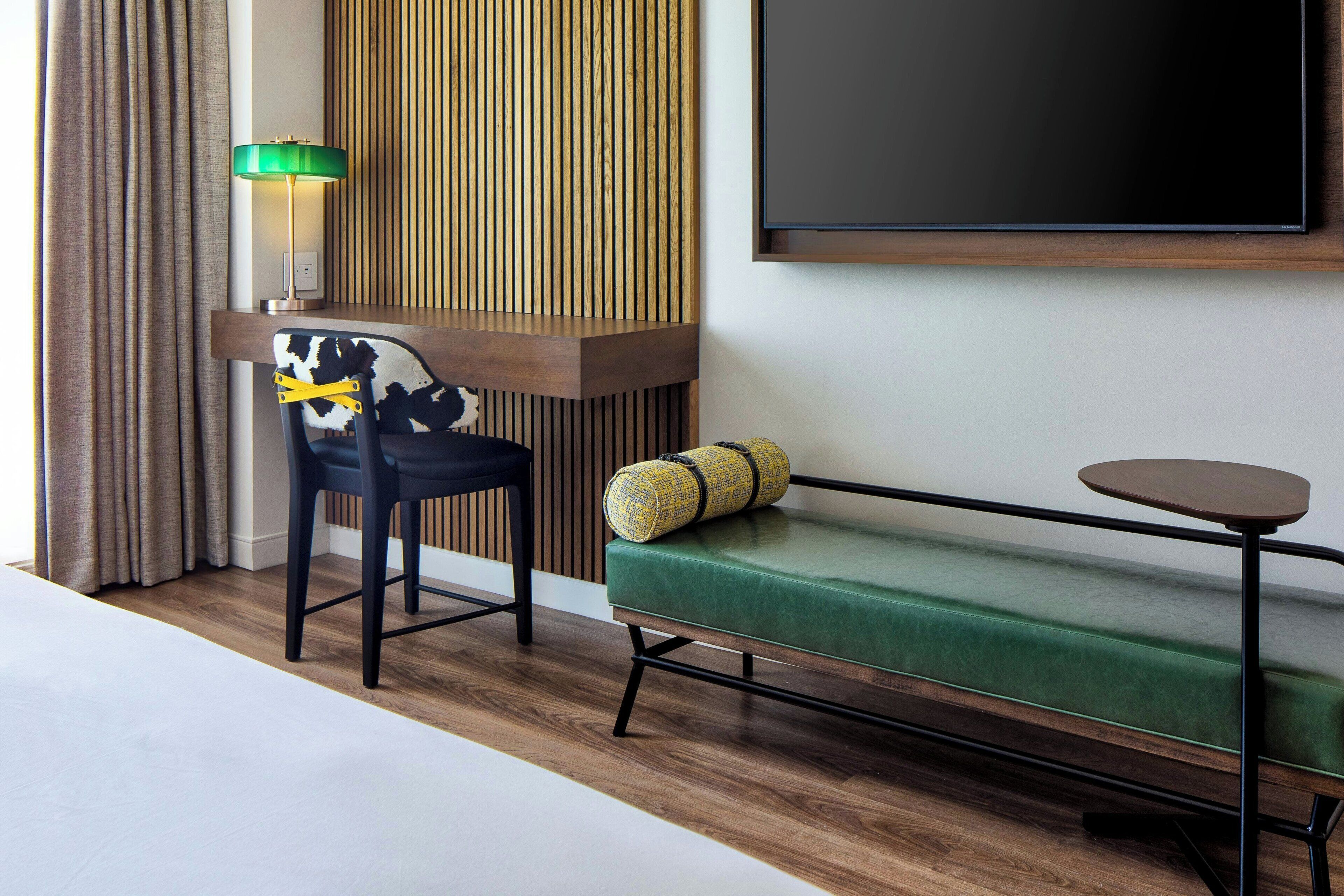 Una habitación de hotel moderna con piso de madera, sofá verde, una mesita redonda pequeña, televisor negro y lámpara con pantalla verde.