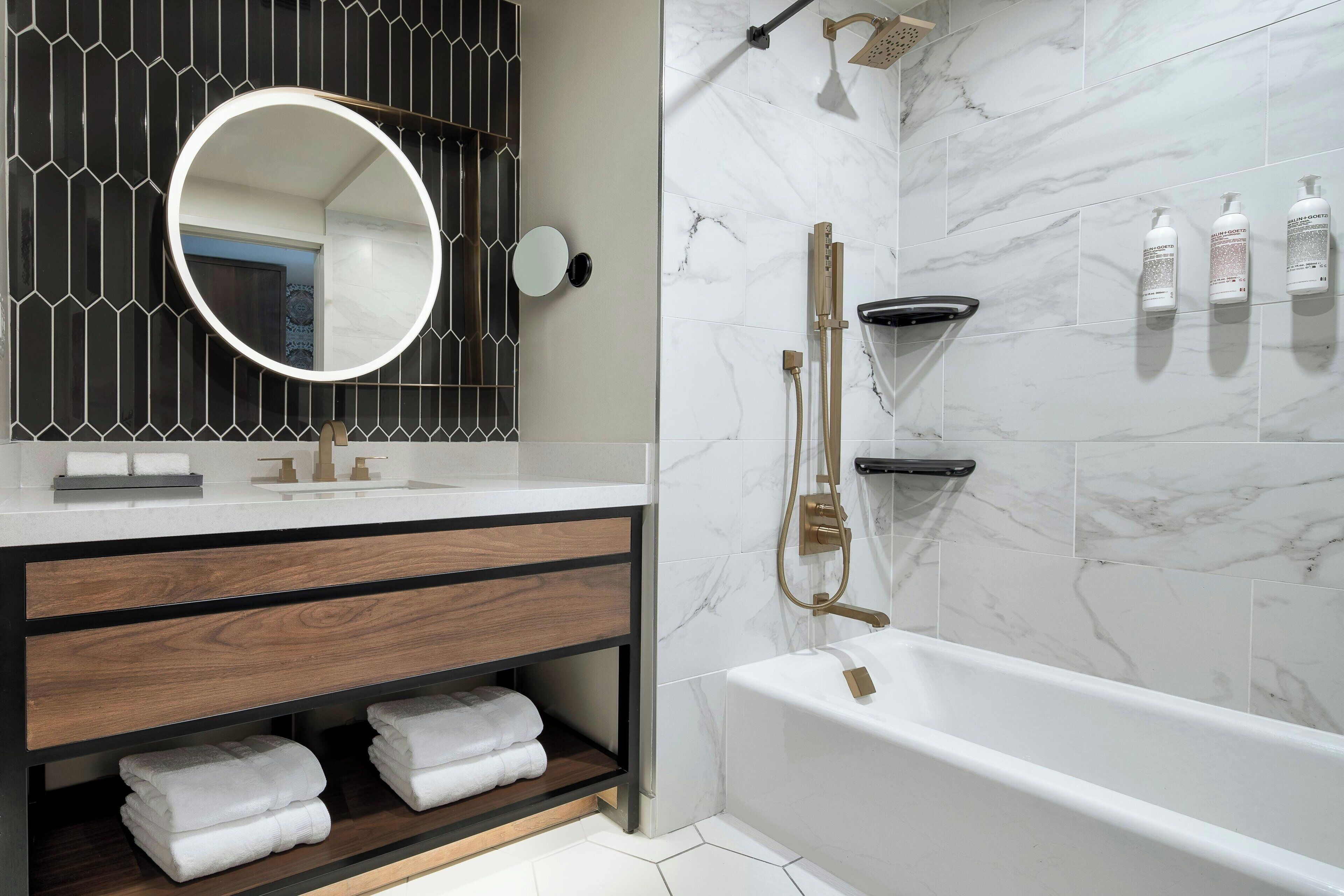 A modern bathroom with a marble shower, a round mirror, and a wooden vanity with towels.