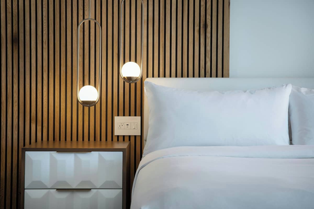 Een netjes opgemaakt bed met witte lakens, een houten lattenpannenhoofd en een nachtkastje met een lade. Twee wandlampen zijn zichtbaar.