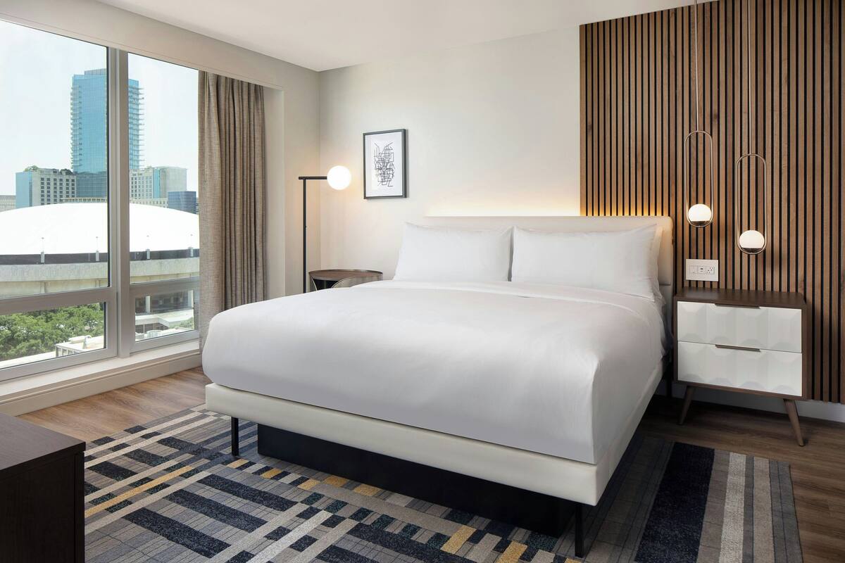 Een moderne hotelkamer met een groot bed, nachtkastje, vloerlamp en uitzicht op de stad.