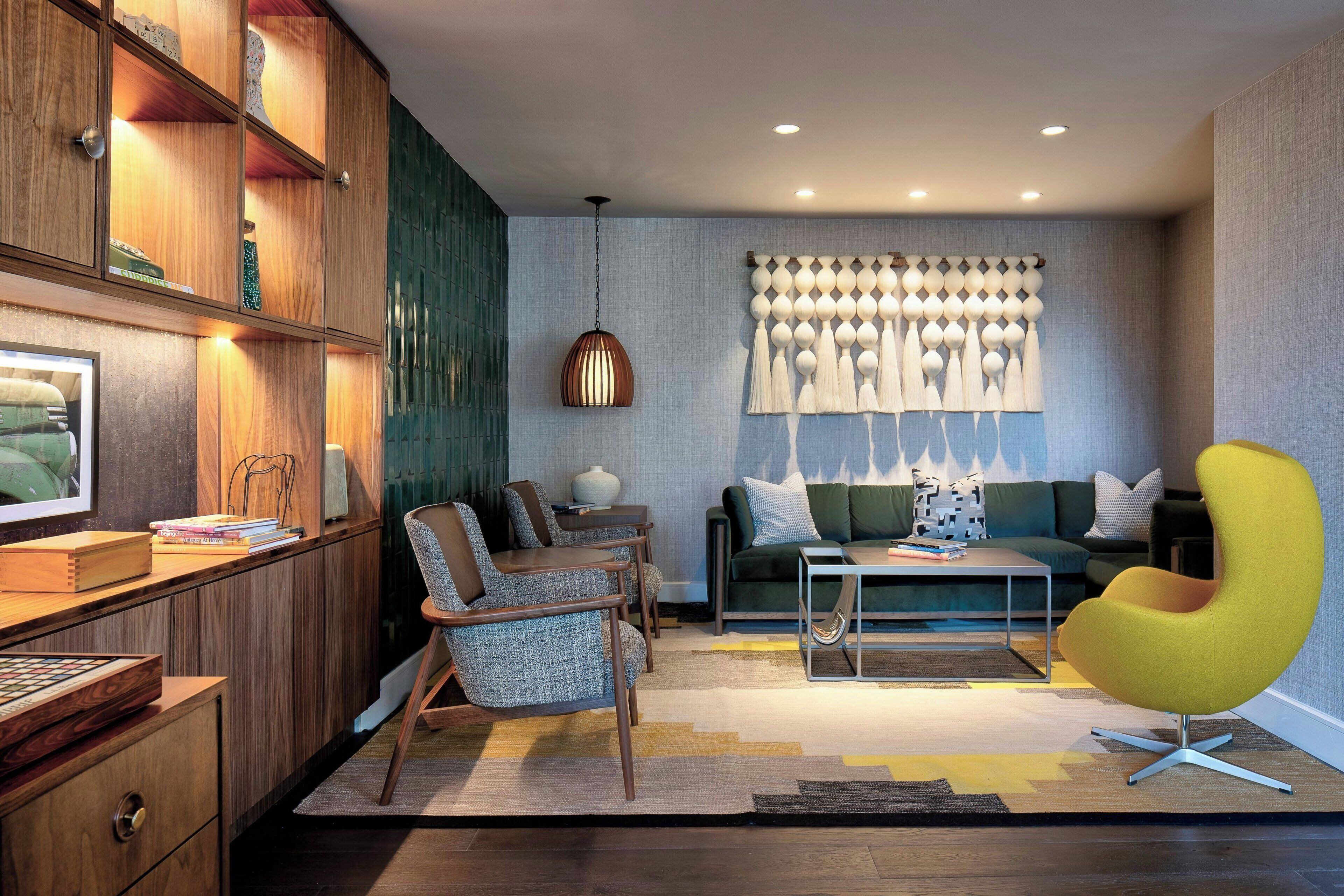 A modern living room with a sofa, chairs, a coffee table, and a yellow accent chair. There is a wooden cabinet with shelves, a hanging light fixture, and a decorative wall panel.