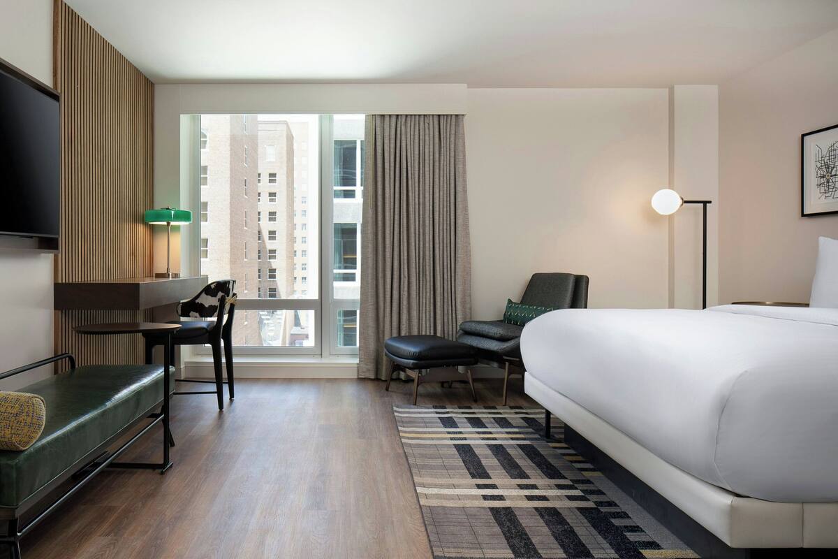 Een hotelkamer met een bed, een bureau, een stoel, een televisie, een lamp en uitzicht op de stad.