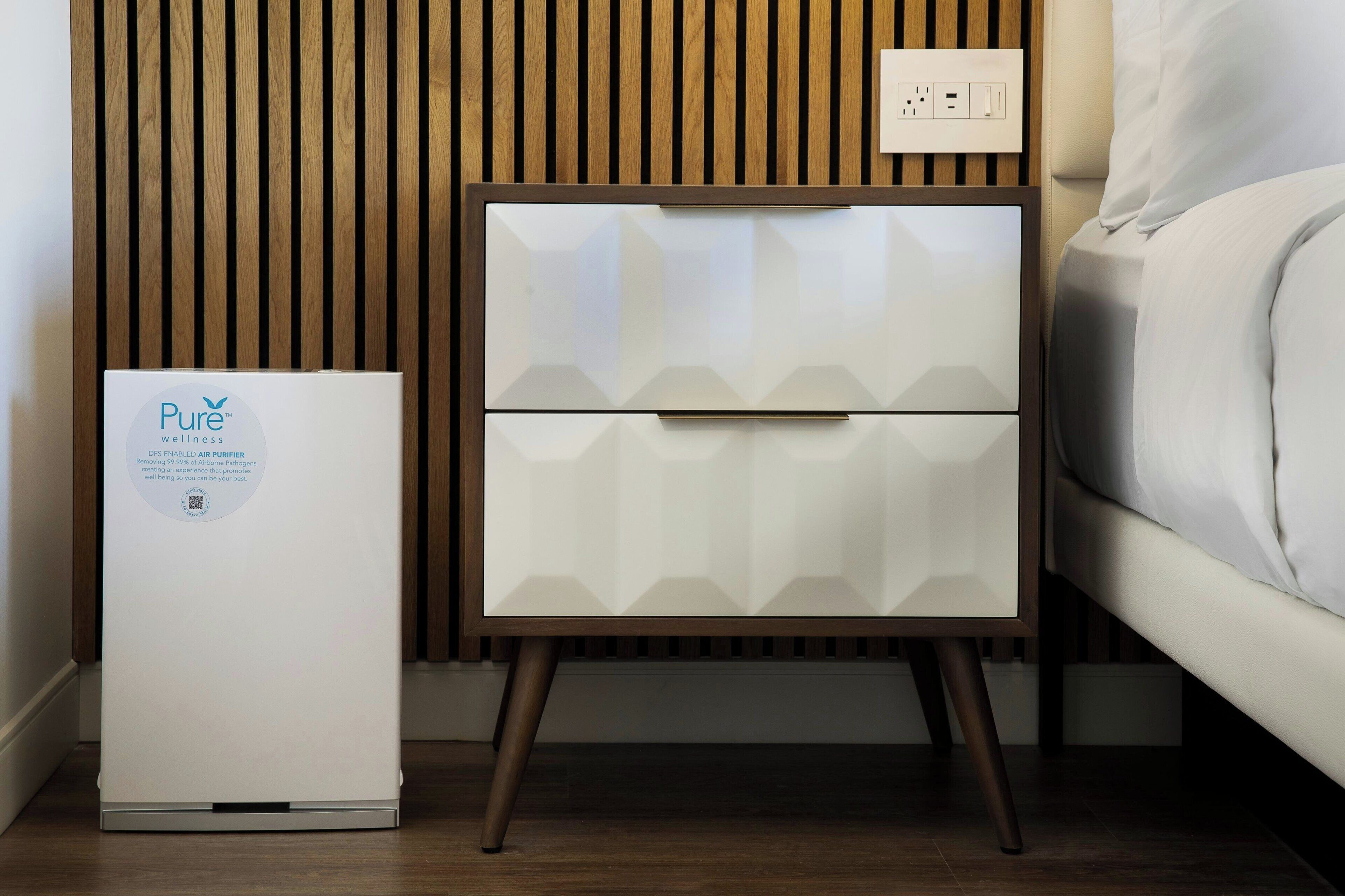 A modern bedroom with a white bed, a nightstand with a white drawer, and a Pure wellness air purifier.