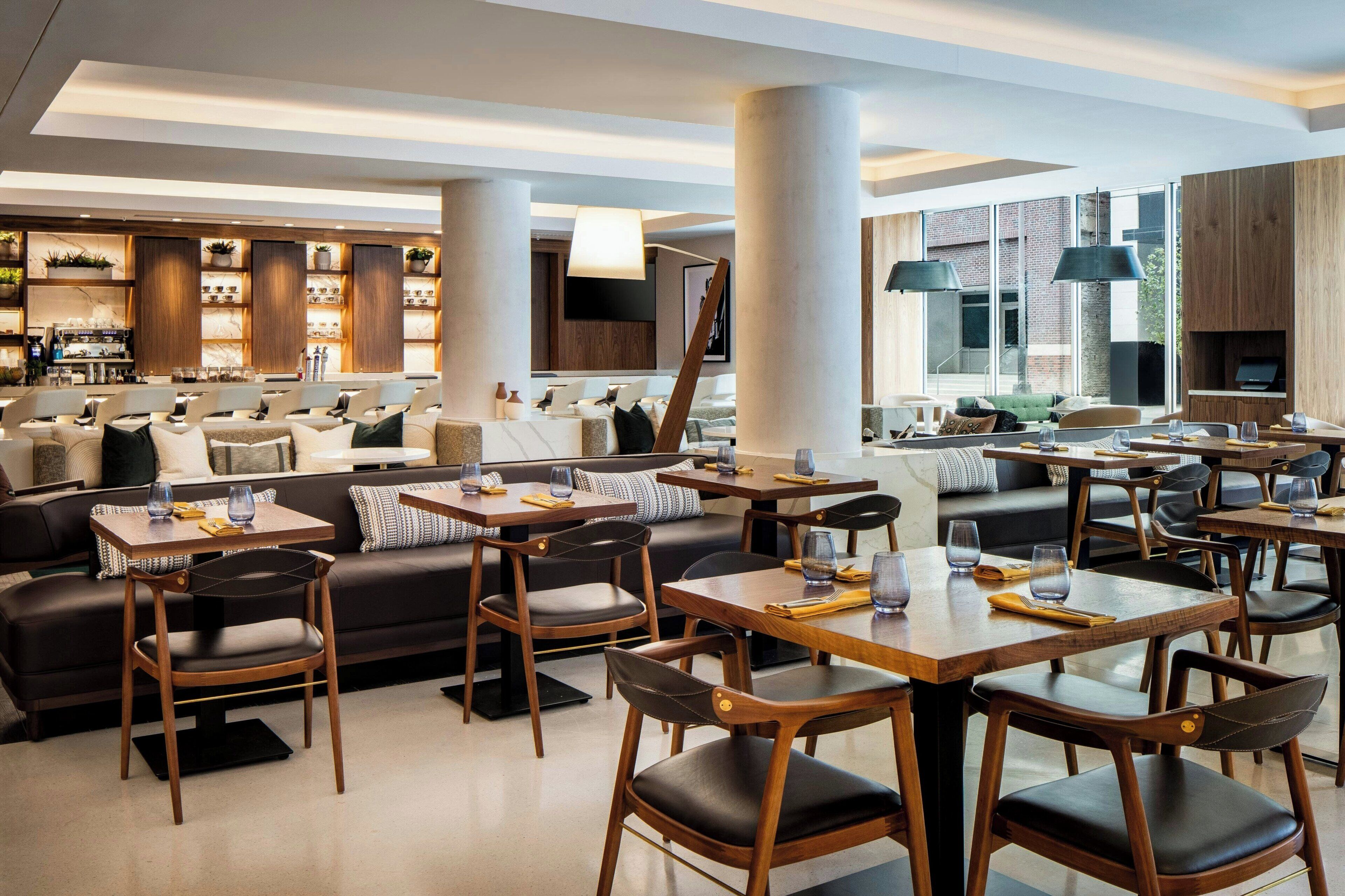 Un restaurante moderno con mesas y sillas de madera, una zona de bar y un rincón de estar acogedor con sofás y cojines.