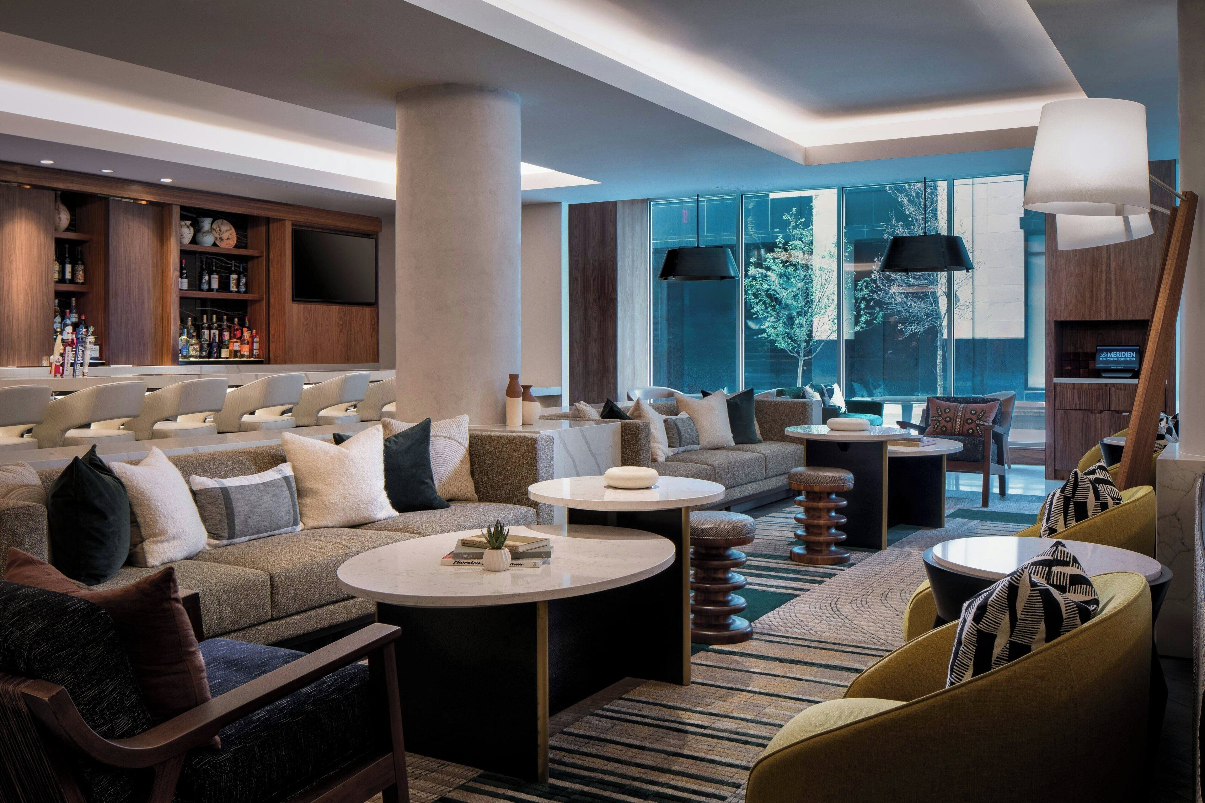 Un moderno vestíbulo de hotel con zona de bar, espacios de descanso y amplios ventanales que permiten la entrada de luz natural.