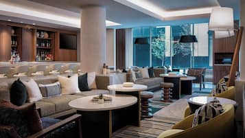 Un moderno vestíbulo de hotel con zona de bar, espacios de descanso y amplios ventanales que permiten la entrada de luz natural.