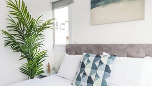 Una cama bien tendida con sábanas blancas, un cabecero tapizado en gris y una almohada con estampado geométrico. Una planta verde grande se encuentra a la izquierda de la cama, y se ve una ventana con vistas a edificios al fondo.