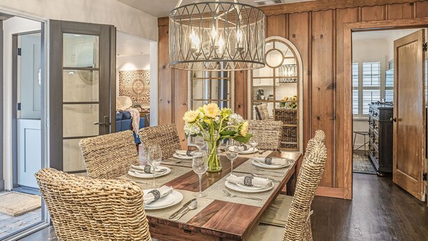 Een eetruimte met een houten tafel voor zes personen, omringd door rieten stoelen, een kroonluchter en een vaas met bloemen.
