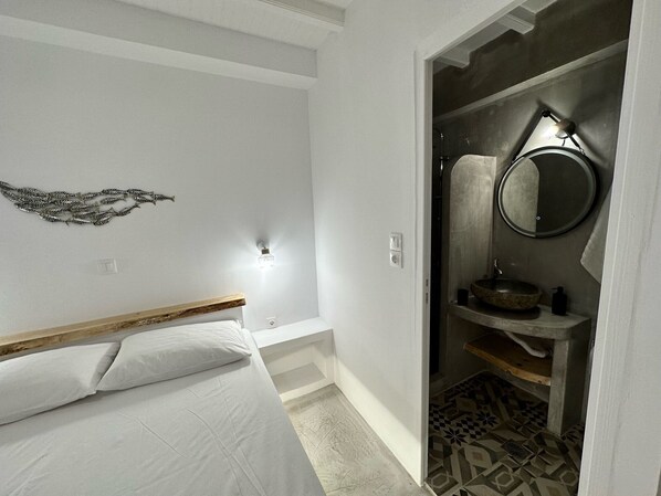 A bedroom with a bed, a wall-mounted light, and a bathroom with a sink and mirror.