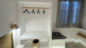 A modern room with a white wall, a wooden headboard, a hanging light fixture, a small bench, a basket, and a window with grey curtains. - Naousa Getaway 2 Bd Dreamy Island Escape (Paros)