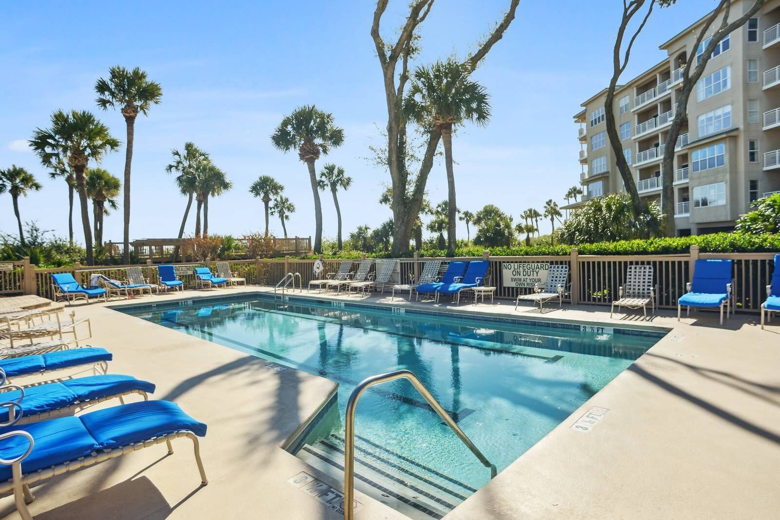 Un espace piscine avec des chaises longues bleues, des palmiers et un bâtiment doté de balcons.