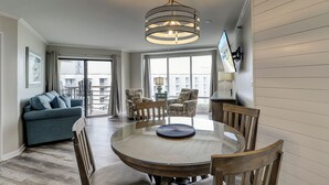 Un salon moderne avec une table ronde en verre, des chaises en bois, un canapé bleu et une grande fenêtre donnant sur un immeuble.