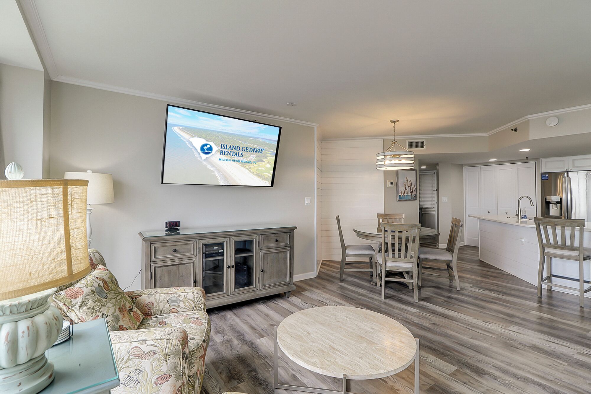 A modern living room with a flat-screen TV, a dining area with wooden chairs, and a kitchen with white cabinets.