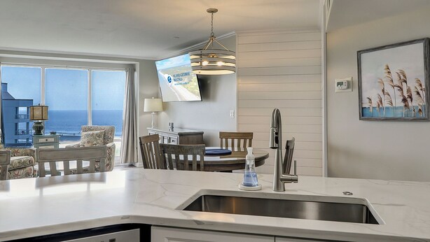 Une cuisine moderne dotée d’une grande fenêtre, un coin repas offrant une vue sur l’océan et un téléviseur fixé au mur qui diffuse des images de « Island Getaway Rentals ».