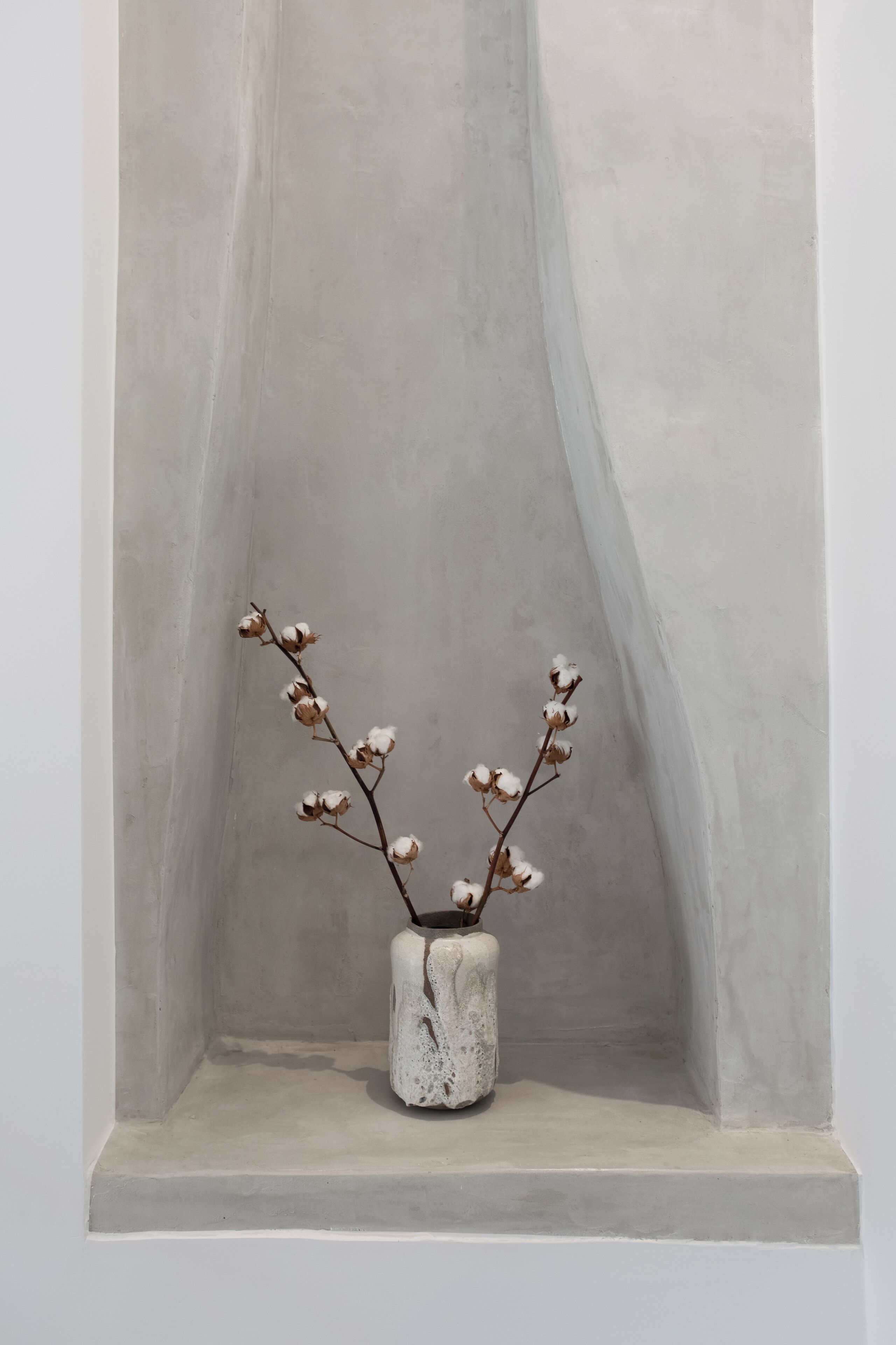 Una jardinera de concreto con una rama de flores de algodón frente a una pared lisa.