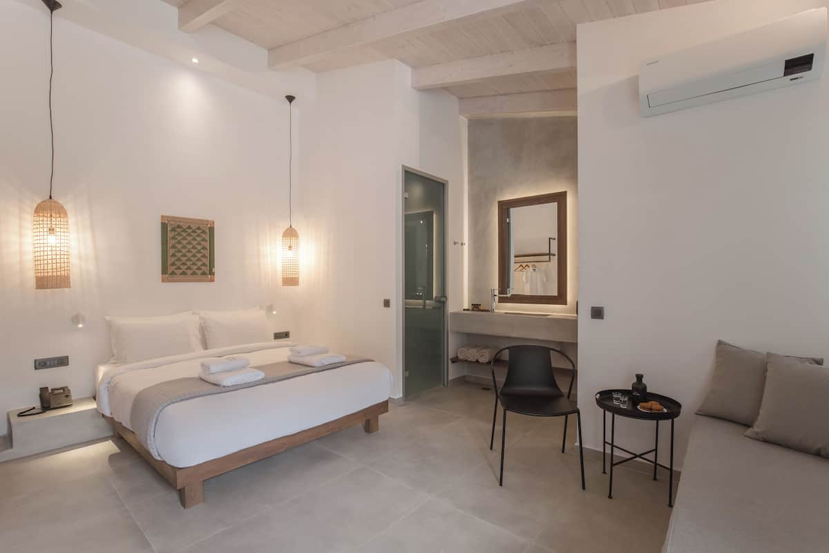 A modern hotel room with a bed, bedside table, chair, and a small table with a cup and a cookie.
