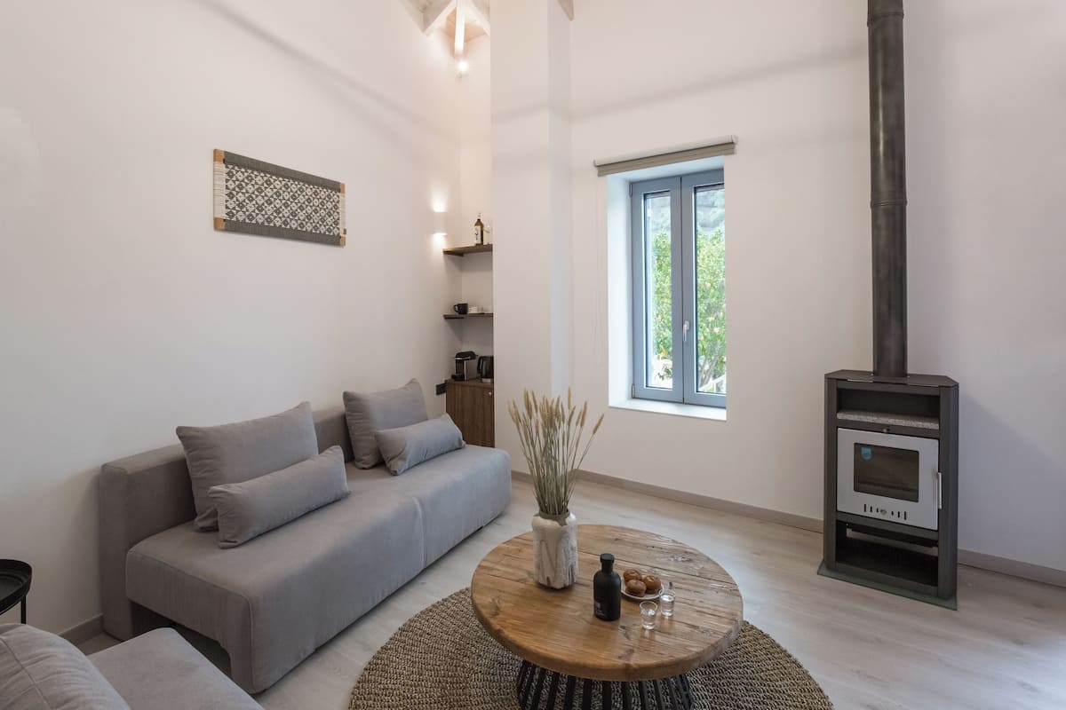 A modern living room with a grey sofa, a round wooden coffee table with decorative items, a built-in wood stove, and a window with a view of greenery.