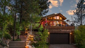 Un chalet rustique avec un escalier en pierre menant à une terrasse en bois, entouré d’arbres et bordé d’une allée en gravier.