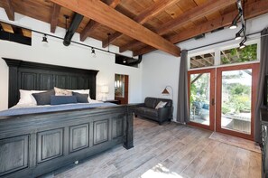 Un dormitorio con una cama grande, un cabecero de madera oscura, una mesita de noche, un escritorio, un sofá de cuero y un ventanal corredera que da a un balcón.