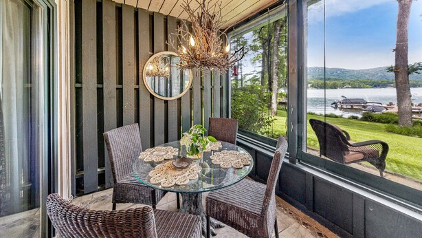 Une table en verre, des chaises en osier, un miroir rond et un lustre dans une pièce dotée d’un plafond en bois et de grandes fenêtres donnant sur un lac.