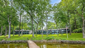 Un pont en bois enjambe un étang et mène à un bâtiment doté de nombreux balcons et d’une pelouse.