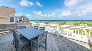 Eine Holzterrasse mit Tisch und Stühlen, mit Blick auf einen Strand und ein Haus.