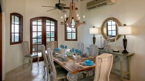 A dining area with a wooden table, chairs, and a chandelier. There are windows with views, a ceiling fan, and an air conditioning unit. - Bananaquit Villa: Amazing Views! Large Pool! Full AC! (St John)